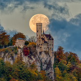Eine Burg auf einem Hügel und einem wolkenbehangenen Himmel. Hinter dem Burgturm ist der Vollmond.