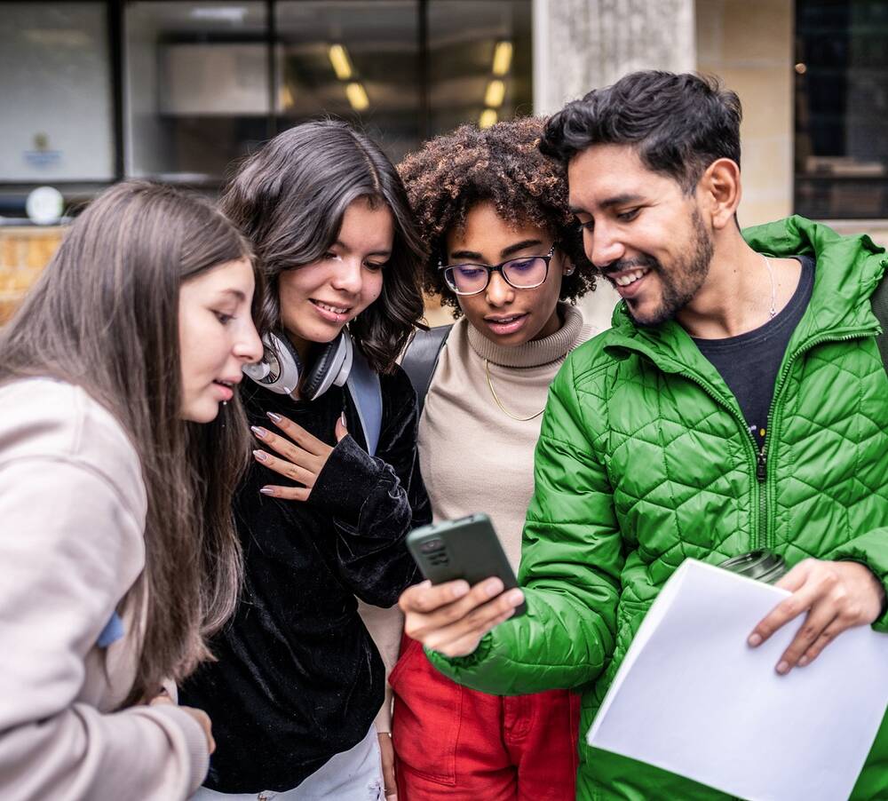 Eine Gruppe von vier jungen Erwachsenen steht eng zusammen und schaut lächelnd auf ein Smartphone, das einer von ihnen in der Hand hält.