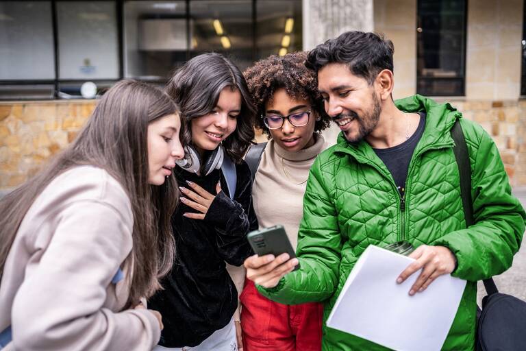 Eine Gruppe von vier jungen Erwachsenen steht eng zusammen und schaut lächelnd auf ein Smartphone, das einer von ihnen in der Hand hält.