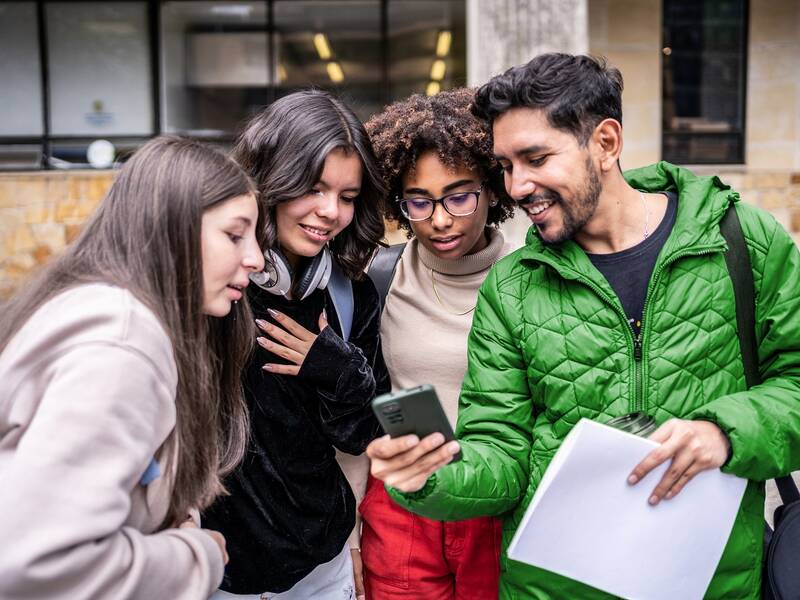 Eine Gruppe von vier jungen Erwachsenen steht eng zusammen und schaut lächelnd auf ein Smartphone, das einer von ihnen in der Hand hält.
