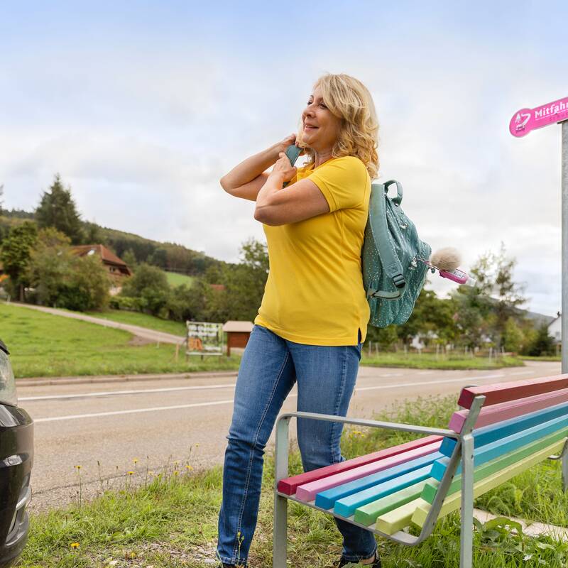 Eine leger gekleidete blonde Frau mit Rucksack steht an einer bunten Bank. Die Bank befindet sich an einer Landstraße.