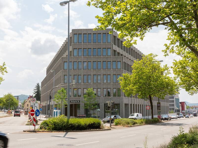 Ein modernes Bürogebäude mit mehreren Etagen und Glasfassade steht an einer belebten Straßenecke, umgeben von Bäumen und Verkehrsschildern.