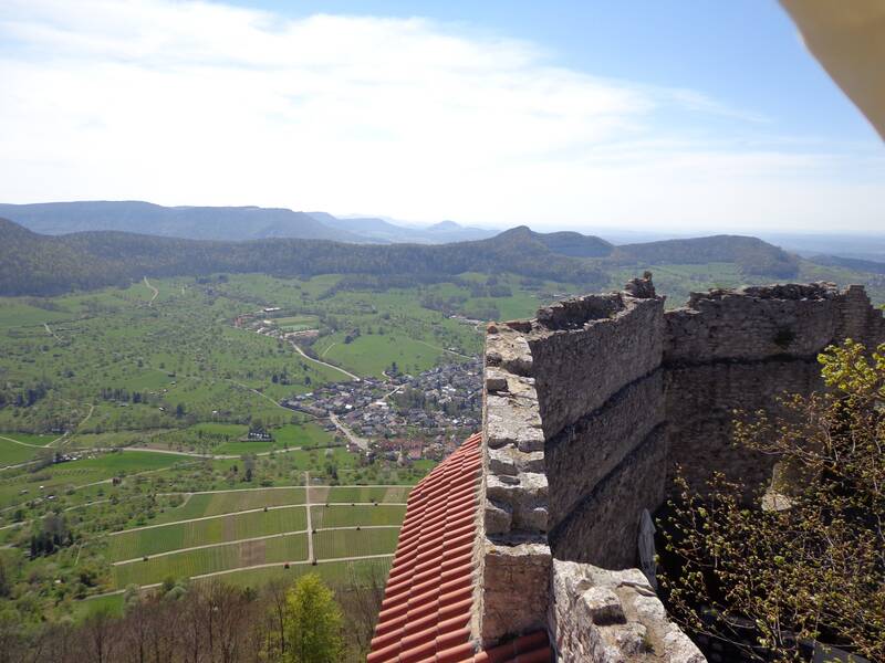 Die Aussicht von einer alten Steinmauer bietet einen weiten Blick über eine grüne Landschaft mit Feldern, einem kleinen Dorf und entfernten Hügeln.