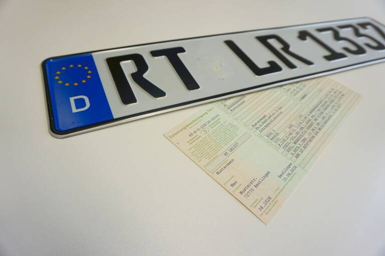 Ein Autokennzeichen mit der Kennzeichnung RT LR 1337 liegt auf einem weißen Tisch. Daneben liegt eine hellgrüne Zulassungsbescheinigung.