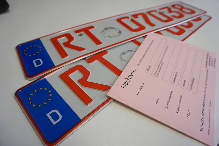 Zwei Autokennzeichen für Odltimer liegen auf einem weißen Tisch. Die Buchstaben und Zahlen auf dem Kennzeichen sind in roter Schrift. Auf dem Kennzeichen liegt ein rosafarbenes Formular.