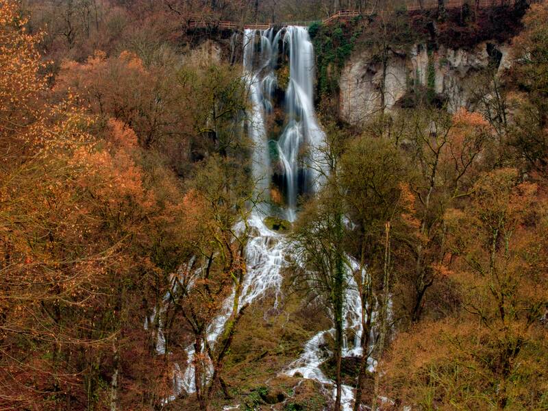 Ein hoher Wasserfall fließt durch eine herbstliche Waldlandschaft mit buntem Laub.