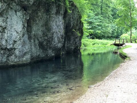 Ein klarer Fluss fließt an einer steilen Felswand vorbei, neben einem schmalen Weg mit einer kleinen Holzbrücke umgeben von grüner Vegetation.
