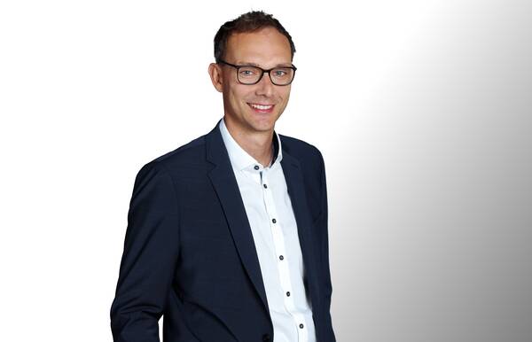 Ein lächelnder Mann mit Brille, gekleidet in ein dunkles Sakko über einem weißen Hemd, steht vor einem hellen Hintergrund.
