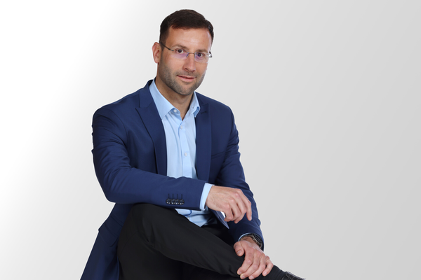 Ein Mann mit Brille und Bart sitzt entspannt mit übergeschlagenen Beinen und trägt einen dunkelblauen Anzug sowie ein hellblaues Hemd vor einem weißen Hintergrund.