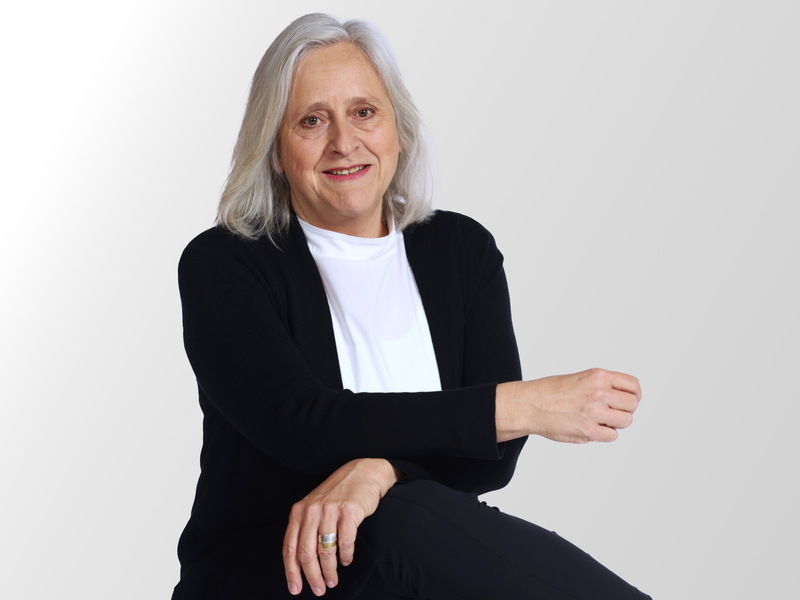Ein Portrait einer Frau mit schulterlangen grauen Haaren. Sie trägt ein weißes Shit mit einem schwarzen Pullover.