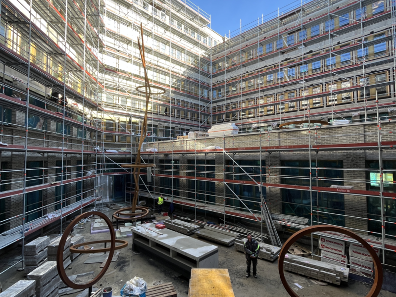 Ein Innenhof eines im Bau befindlichen Gebäudes ist mit Gerüsten umgeben, auf dem Boden liegen Baumaterialien und blaue Müllsäcke, und es gibt zwei große runde Metallstrukturen.