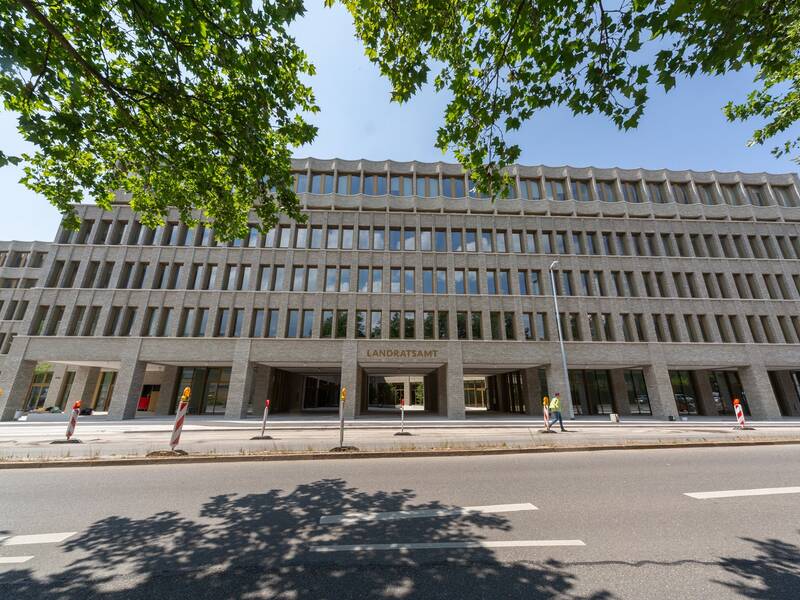 Das Bild zeigt ein modernes, mehrstöckiges Gebäude mit symmetrischen Fensterreihen und einem Eingangsbereich, der von Bäumen und einer Straße im Vordergrund umgeben ist.
