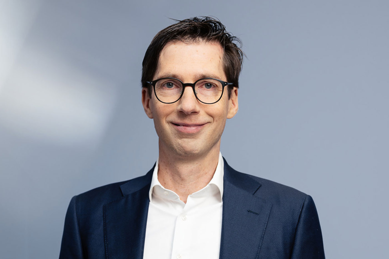 Ein Mann mit Brille, in einem dunklen Anzug und weißem Hemd, lächelt vor einem grau-blauen Hintergrund.
