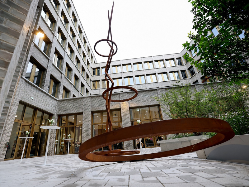 In einem Innenhof mit umliegenden Bürogebäuden steht eine moderne Skulptur mit spiralförmigen Metallringen auf einem gepflasterten Boden.