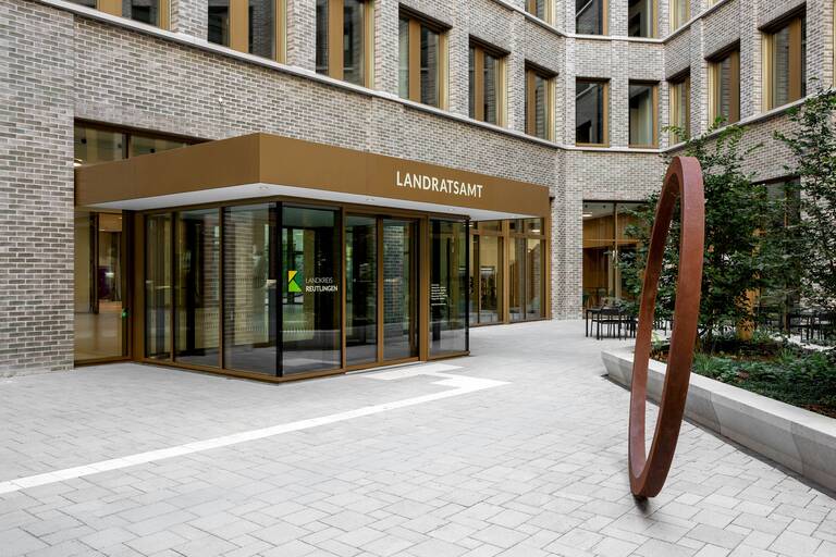 Eingang zu einem modernen Landratsamt mit einer Glasfront, umgeben von Backsteinwänden, und einer großen, metallenen Skulptur im Innenhof.
