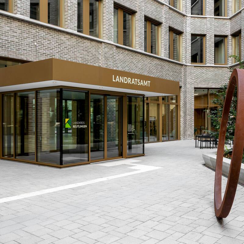 Eingang zu einem modernen Landratsamt mit einer Glasfront, umgeben von Backsteinwänden, und einer großen, metallenen Skulptur im Innenhof.