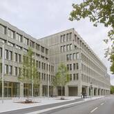 Ein großes, modernes Gebäude aus grauem Backstein mit vielen Fenstern steht an einer Straßenecke, umgeben von jungen Bäumen und Bürgersteigen.