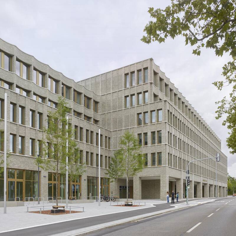 Ein großes, modernes Gebäude aus grauem Backstein mit vielen Fenstern steht an einer Straßenecke, umgeben von jungen Bäumen und Bürgersteigen.