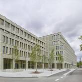Ein modernes, mehrstöckiges Gebäude mit einer Fassade aus hellen Ziegelsteinen steht an einer Straßenecke, umgeben von einigen Bäumen und einem Gehweg.