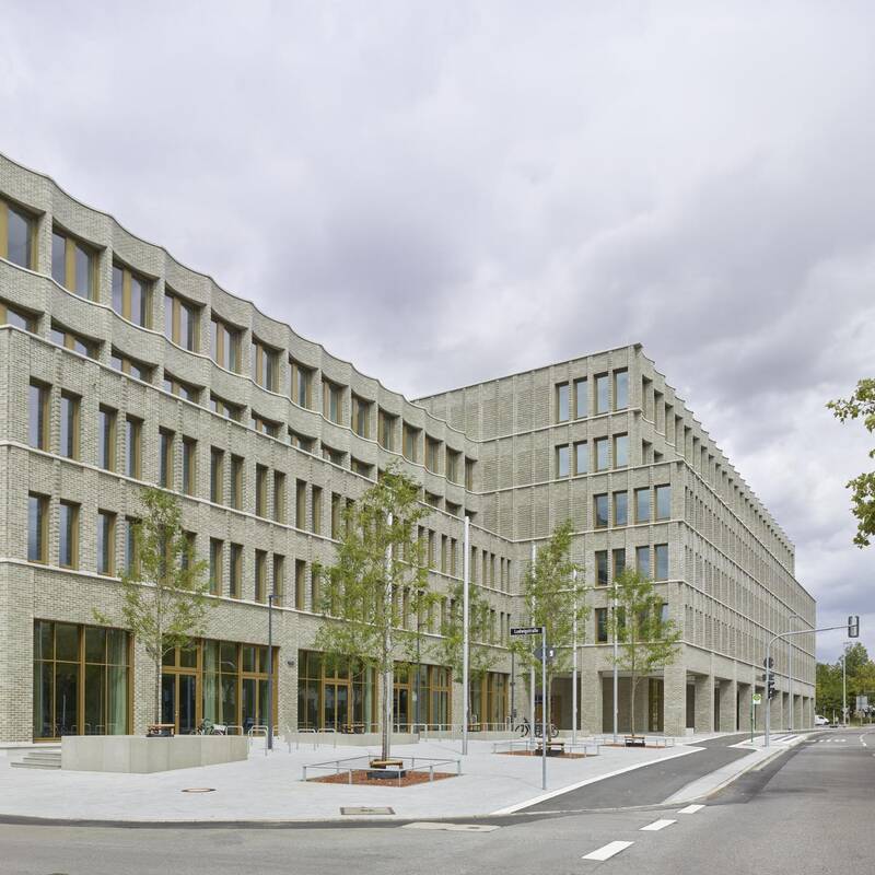 Ein modernes, mehrstöckiges Gebäude mit einer Fassade aus hellen Ziegelsteinen steht an einer Straßenecke, umgeben von einigen Bäumen und einem Gehweg.