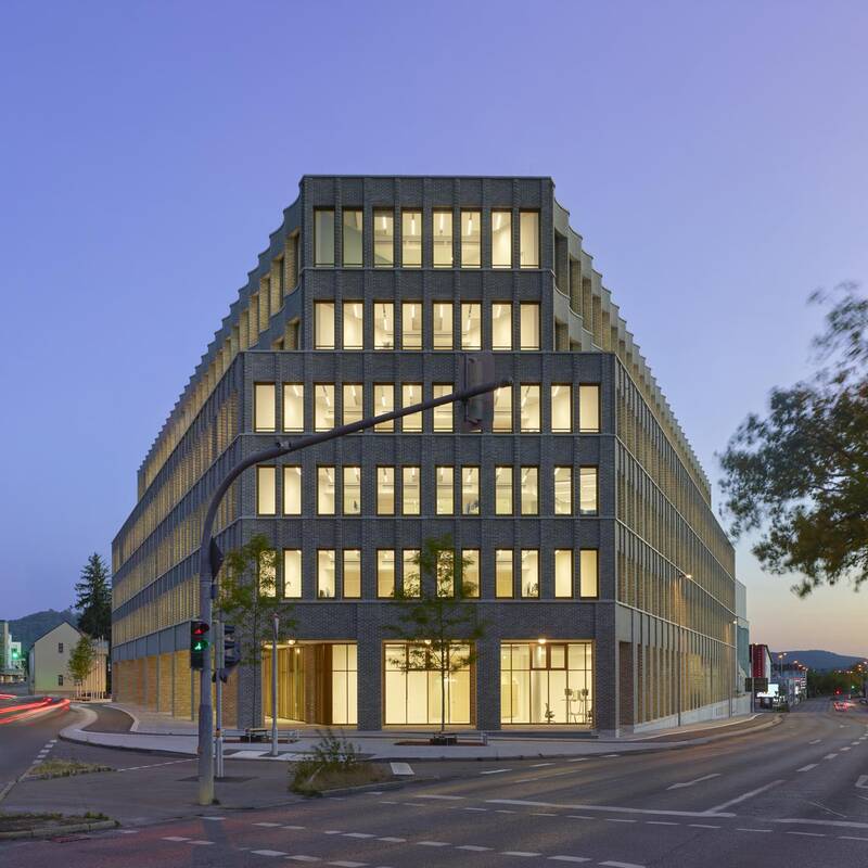 Ein modernes, fünfstöckiges Bürogebäude mit beleuchteten Fenstern steht an einer Straßenkreuzung in der Dämmerung.
