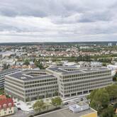 Ein modernes, großes Gebäude mit Solarpanelen auf dem Dach steht inmitten einer Stadtlandschaft unter bewölktem Himmel.