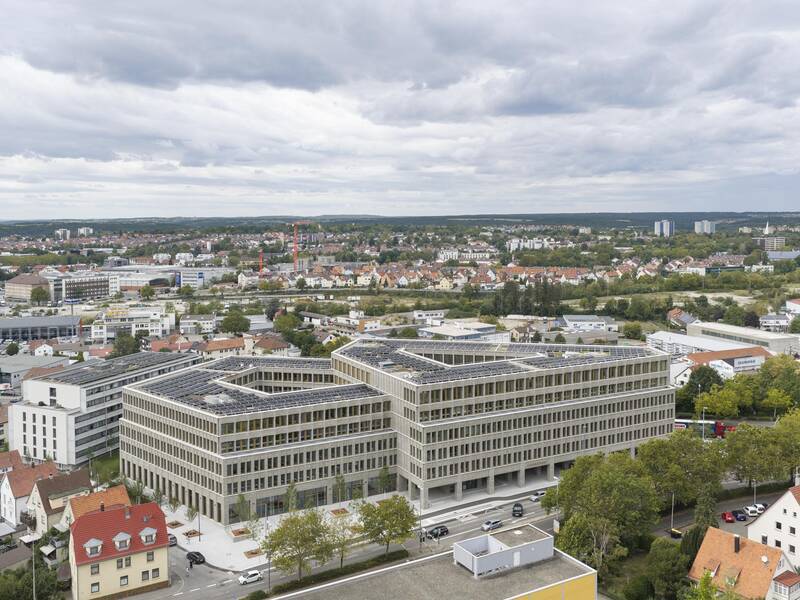 Ein modernes, großes Gebäude mit Solarpanelen auf dem Dach steht inmitten einer Stadtlandschaft unter bewölktem Himmel.