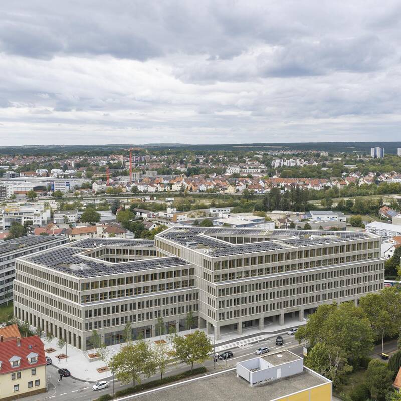 Ein modernes, großes Gebäude mit Solarpanelen auf dem Dach steht inmitten einer Stadtlandschaft unter bewölktem Himmel.
