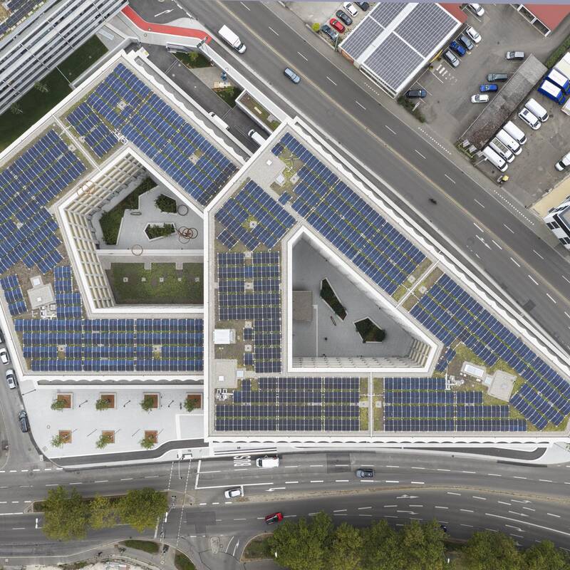 Das Bild zeigt ein dreieckiges Gebäude aus der Vogelperspektive, dessen Dach vollständig mit Solarmodulen bedeckt ist, umgeben von Straßen und weiteren Gebäuden.
