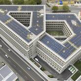 Das Bild zeigt ein großes, modernes Gebäude mit V-förmiger Struktur, das mit zahlreichen Solarpaneelen auf dem Dach bedeckt ist und von mehreren Straßen umgeben ist.