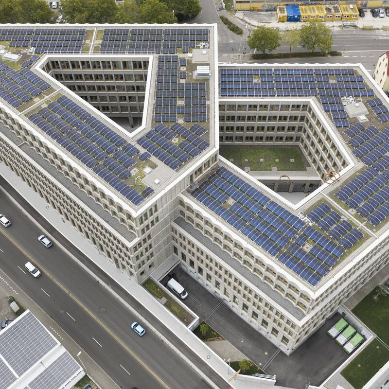 Das Bild zeigt ein großes, modernes Gebäude mit V-förmiger Struktur, das mit zahlreichen Solarpaneelen auf dem Dach bedeckt ist und von mehreren Straßen umgeben ist.