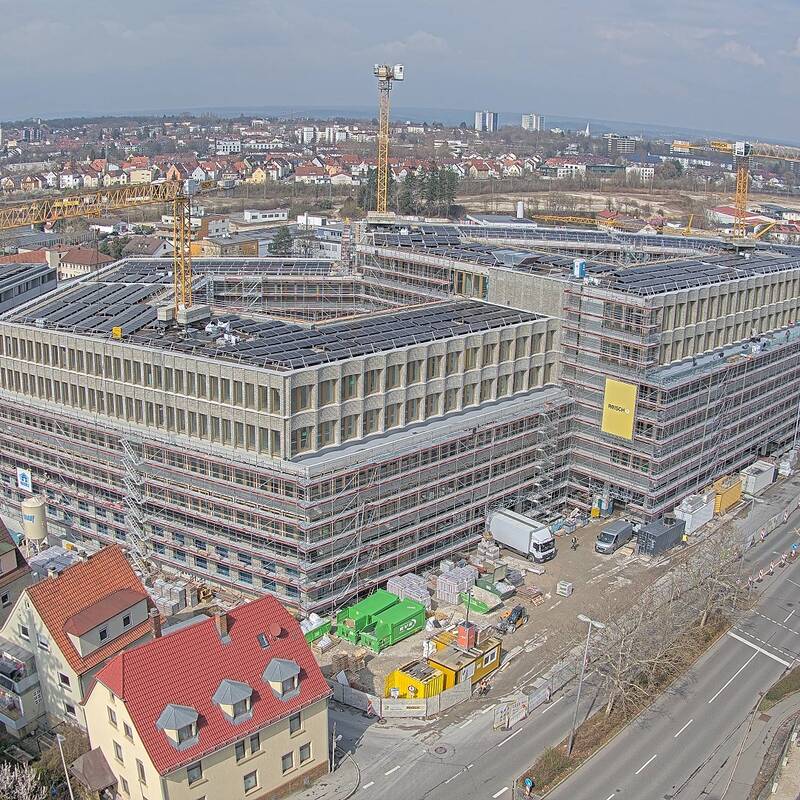 Eine große Baustelle mit mehreren Gebäuden in verschiedenen Bauphasen, von Gerüsten umgeben, in einer städtischen Umgebung mit Krähnen im Hintergrund.