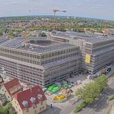 Ein großes, modernes Gebäude ist von einem Baugerüst umgeben, während zwei Baukräne darüber ragen, inmitten einer städtischen Umgebung mit Wohnhäusern und Bäumen.