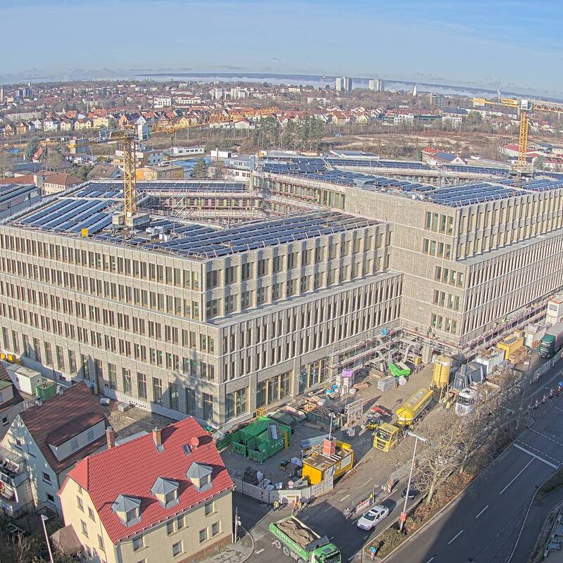 Das Bild zeigt eine große Baustelle mit mehreren Kränen, umgeben von städtischer Infrastruktur und Wohngebäuden.