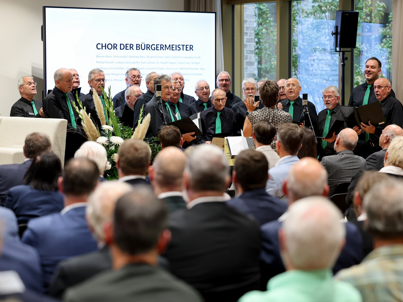 Eine Gruppe von Männern singt als Chor vor einem Publikum, geleitet von einer Dirigentin, mit einer Präsentation im Hintergrund, die "Chor der Bürgermeister" anzeigt.