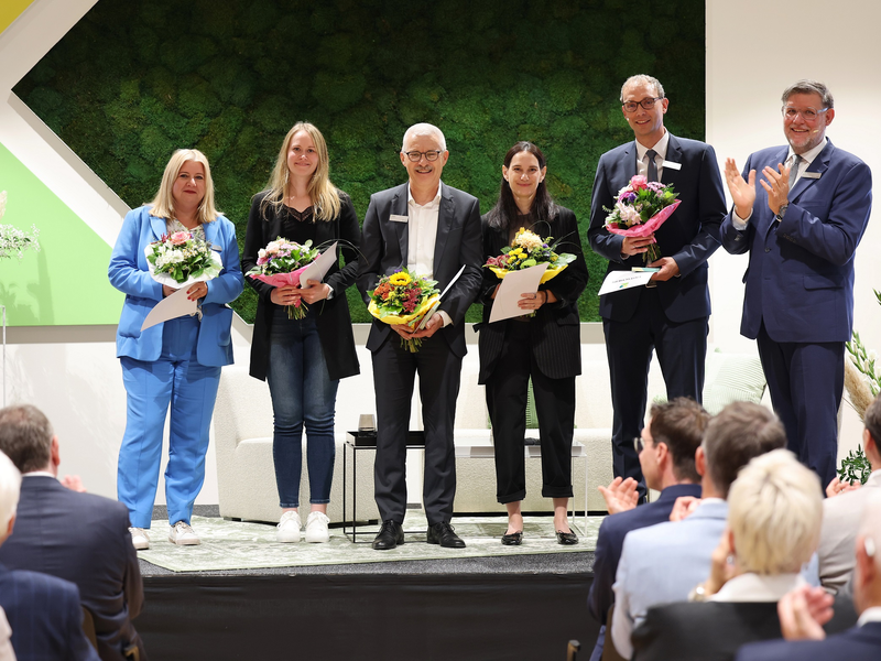 Sechs Menschen in formeller Kleidung stehen nebeneinander auf einer Bühne, halten Blumensträuße und Urkunden in den Händen, während ein Publikum applaudiert.