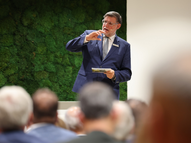 Ein Mann im Anzug hält eine Rede vor einem Publikum und gestikuliert mit der Hand, während er ein Blatt Papier hält, vor einem Hintergrund aus grünem Moos.