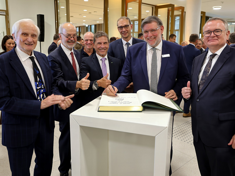 Eine Gruppe von Männern in Anzügen steht um einen Tisch, auf dem ein großes Buch liegt; einer von ihnen schreibt darin, während andere Daumen hoch zeigen.