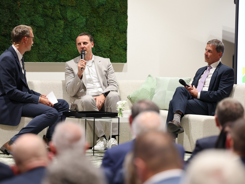 Drei Männer in Anzügen sitzen zusammen auf einem Podium und halten Mikrofone, während sie vor einem Publikum diskutieren.