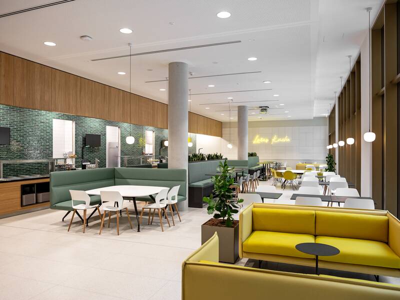 Ein moderner, offener Cafeteria-Bereich mit grünen und gelben Sitzgelegenheiten, Tischen, Pflanzen und einer leuchtenden Schrift an der Wand.