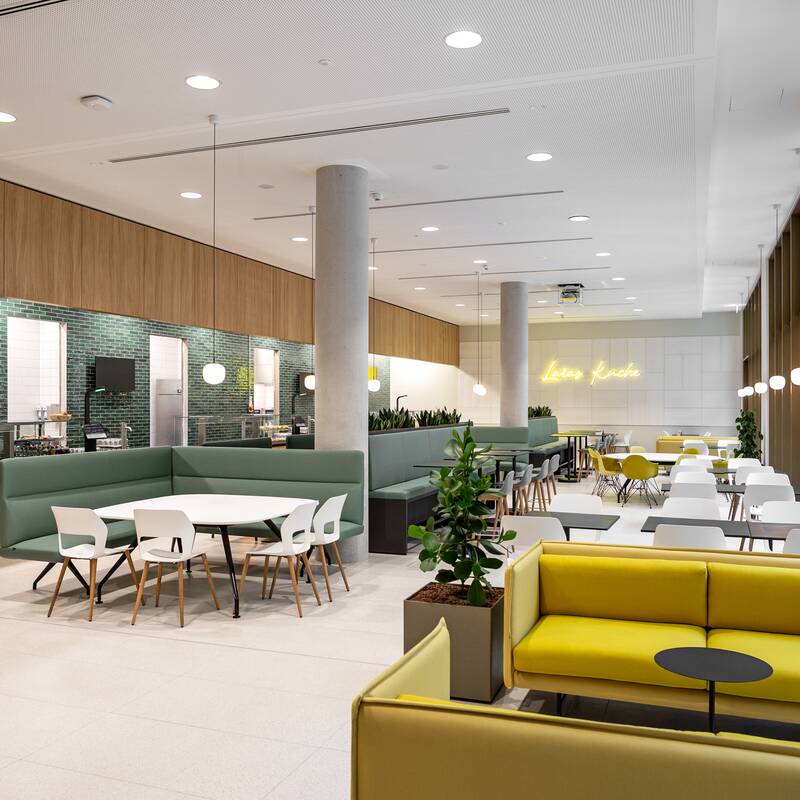 Ein moderner, offener Cafeteria-Bereich mit grünen und gelben Sitzgelegenheiten, Tischen, Pflanzen und einer leuchtenden Schrift an der Wand.
