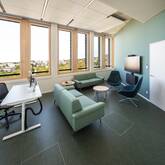 Ein geräumiges, modernes Büro mit großen Fenstern, einer Sitzecke mit Sofas und Sesseln, einem Schreibtisch und einem großen Bildschirm an der Wand.