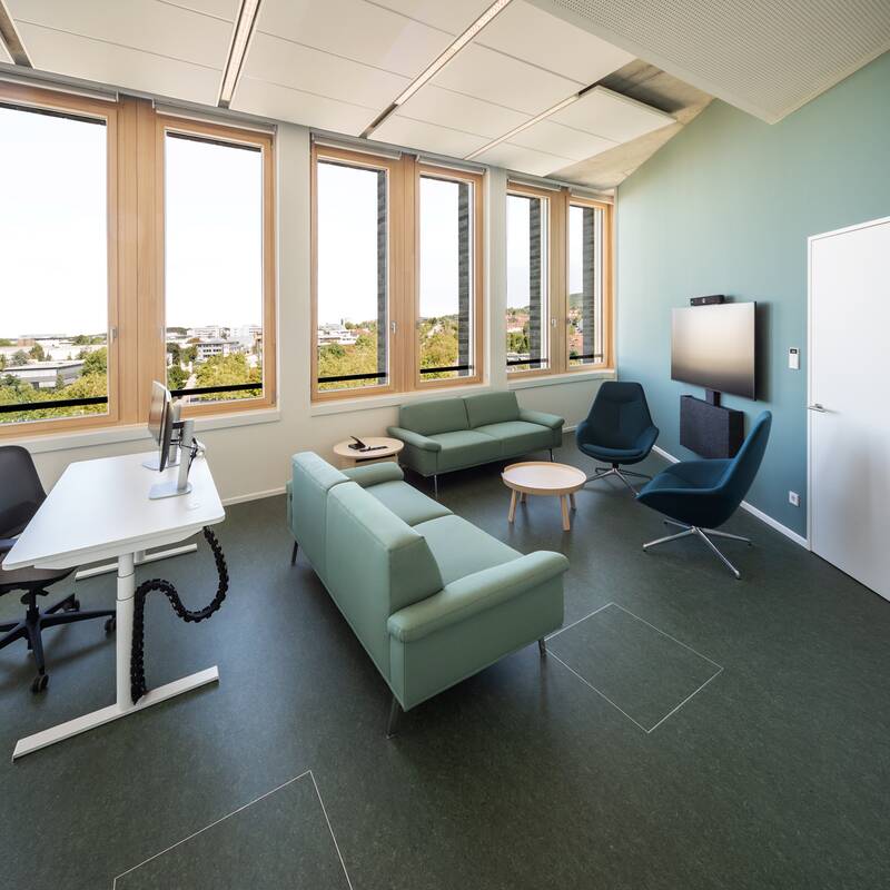 Ein geräumiges, modernes Büro mit großen Fenstern, einer Sitzecke mit Sofas und Sesseln, einem Schreibtisch und einem großen Bildschirm an der Wand.