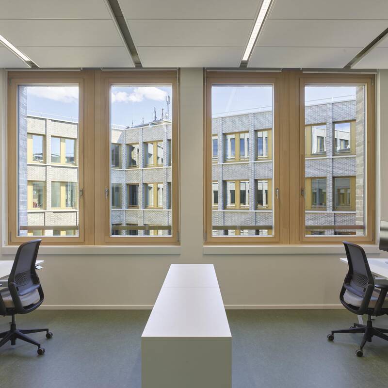 Ein modernes Büro mit zwei Arbeitsplätzen, Schreibtischen und Stühlen, das große Fenster mit Blick auf das angrenzende Gebäude hat.