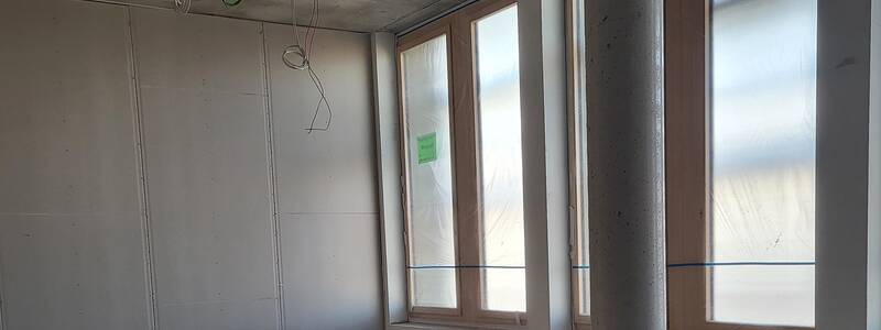 Ein unfertiger Raum mit Betonwänden und -decke, hervorstehenden Kabeln und großen, teils abgedeckten Fenstern.