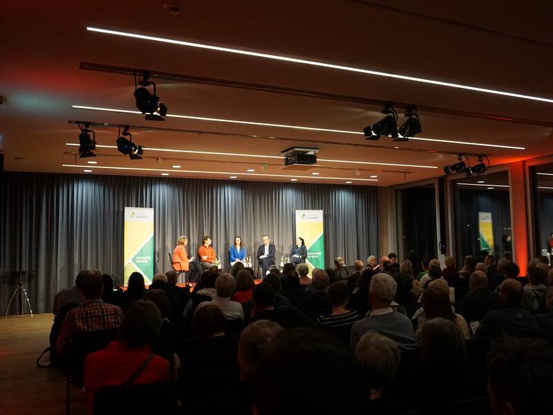 In einem Konferenzraum sitzen mehrere Personen auf einer Bühne vor einem Publikum, wobei der Raum mit Vorhängen und Beleuchtung ausgestattet ist.